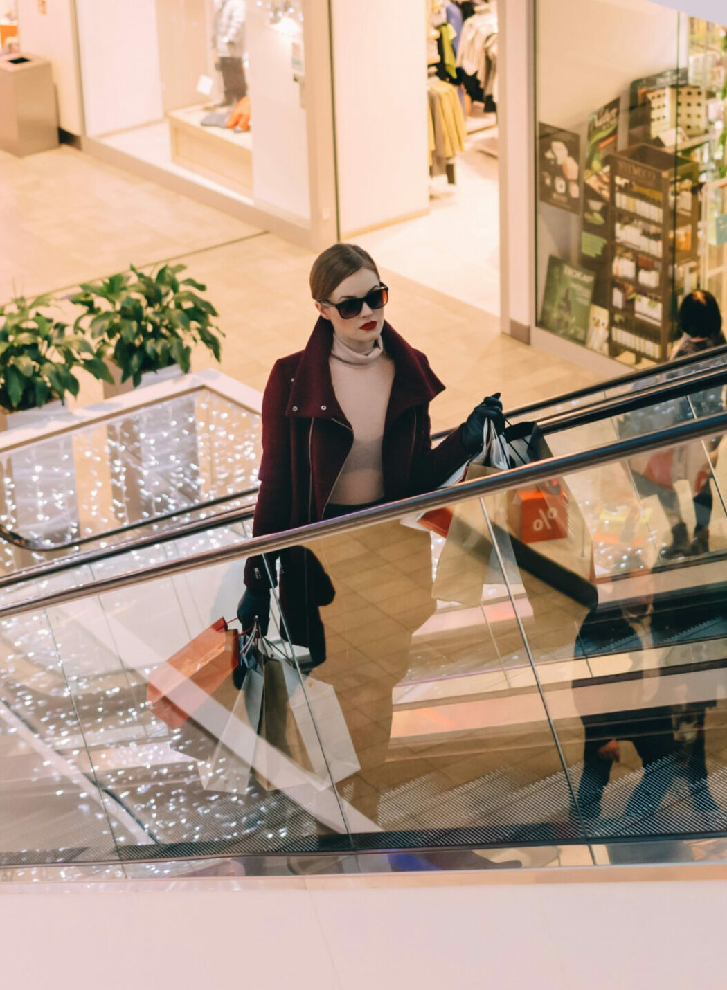 Doftmarknadsföring. En kvinna klädd i en vinröd kappa och solglasögon åker upp för en rulltrappa i ett köpcentrum, med shoppingpåsar i händerna. Bakgrunden visar butiker och julbelysning.