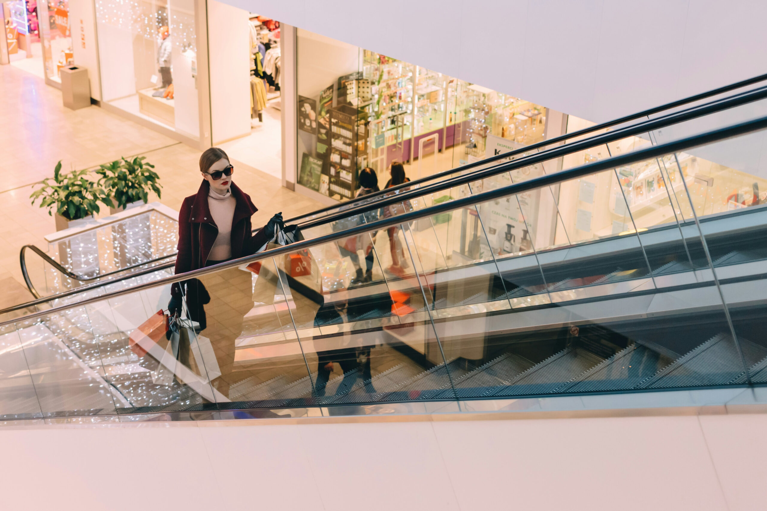 Doftmarknadsföring. En kvinna klädd i en vinröd kappa och solglasögon åker upp för en rulltrappa i ett köpcentrum, med shoppingpåsar i händerna. Bakgrunden visar butiker och julbelysning.